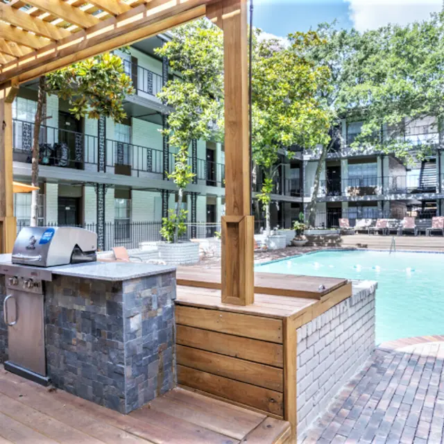 A modern outdoor space featuring a swimming pool with lounge chairs, surrounded by greenery and balconies of an apartment complex. There is a barbecue grill area under a wooden pergola.