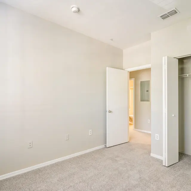 A light-colored, empty bedroom with carpeted floor and two open doors leading to a closet and a bathroom.