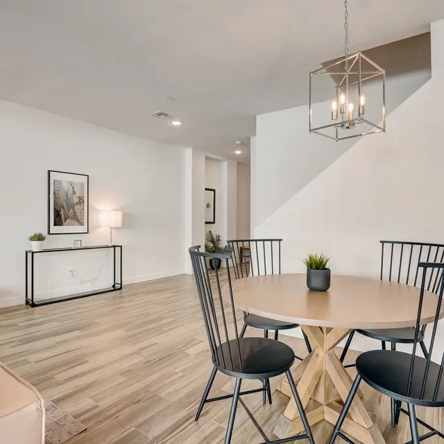 Stylish Modern Dining Area A modern dining area featuring a round wooden table surrounded by black chairs, with a decorative plant on the table. The walls are painted white, and there is a geometric light fixture above. In the background, a cozy seating area with a beige sofa and dark cushions is visible, as well as a piece of artwork on the wall.