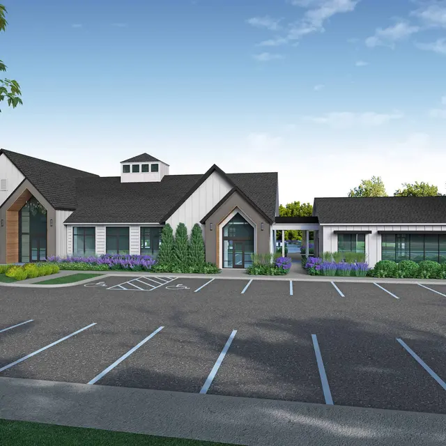 A modern building with a pitched roof and large windows, set in a landscaped area with a parking lot in front. There are trees and shrubs around, with lavender plants visible.