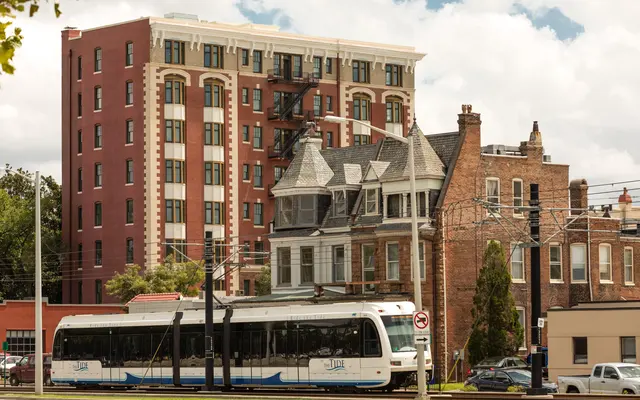 Tram in Urban Landscape A modern tram passes in front of a combination of older and newer buildings. The scene showcases a mix of architectural styles, with a charming Victorian-style house in the foreground and a modern apartment building behind it under a partly cloudy sky.