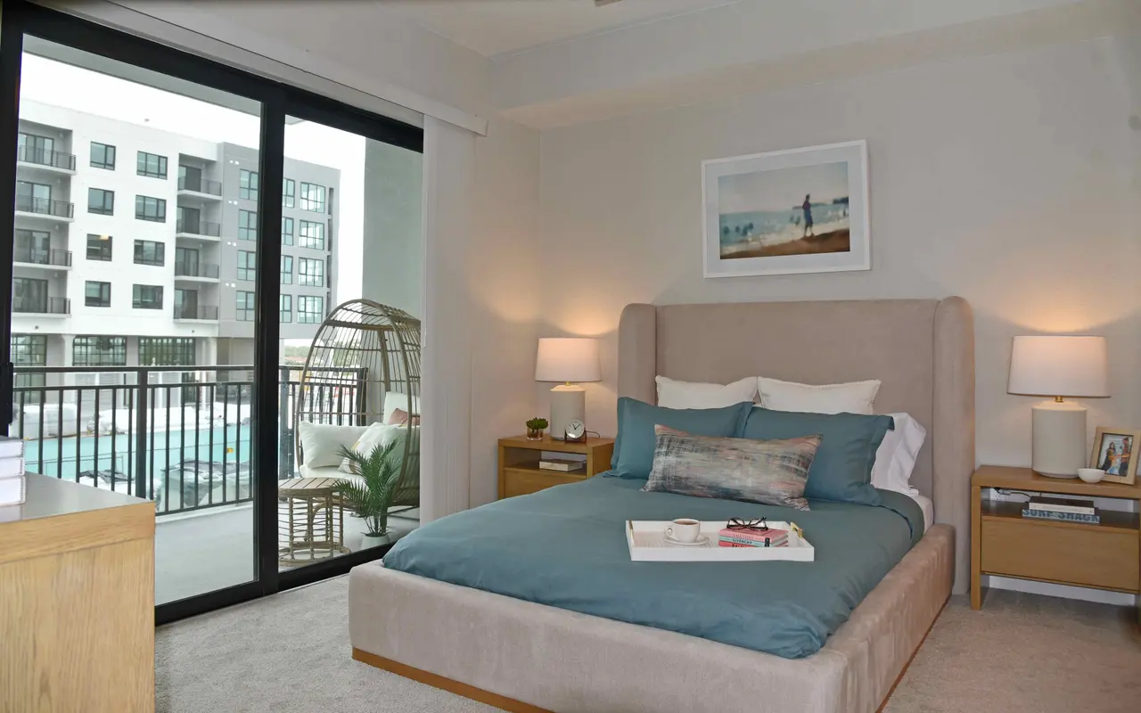 A modern bedroom featuring a large bed with a turquoise blanket, two bedside tables with lamps, and a sliding glass door leading to a balcony. The room is decorated in neutral tones with a framed artwork on the wall.