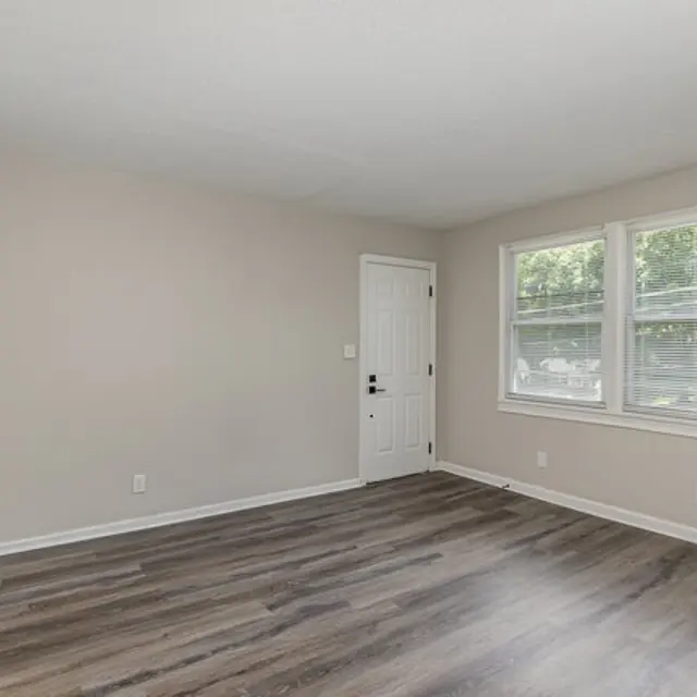 A spacious empty room with a light-colored wall and laminate flooring. There is a door on the left and two windows allowing natural light to flood the space. The room has a clean, modern appearance with no furniture.