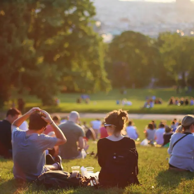 A group of people sitting on a grassy hill during sunset, with some enjoying the view and others engaged in conversation. In the background, more people can be seen in the park enjoying the scenery.