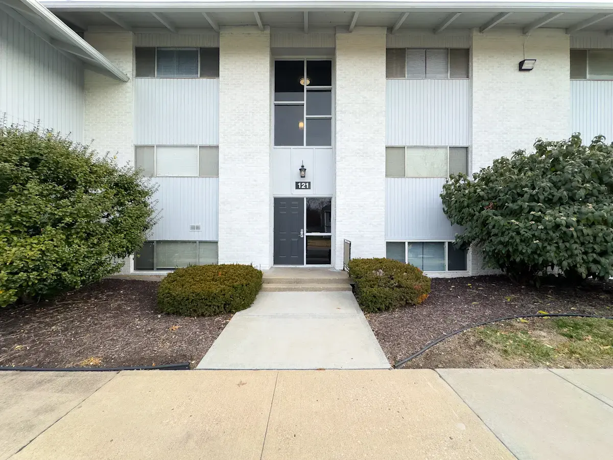 Front view of an apartment building with a clear pathway leading to the entrance, flanked by shrubs and landscaping.