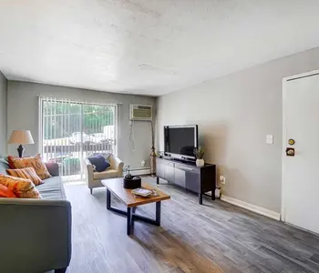 A cozy living room featuring a light gray wall, a blue couch with colorful cushions, a wooden coffee table in the center, and a television on a low cabinet. A lamp stands beside the couch and glass doors lead to an outdoor area.