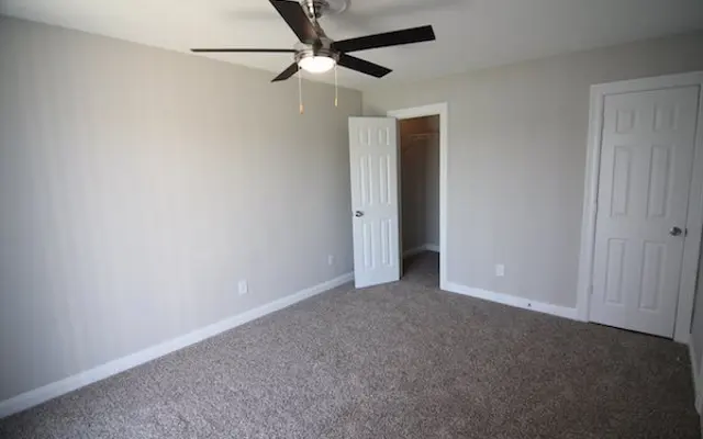Bright Empty Room with Ceiling Fan A spacious, empty room with light gray walls and carpet, featuring a ceiling fan and two doors—one open to a closet. The space is bright and well-lit.