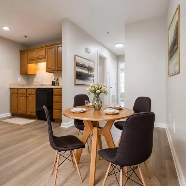 A cozy dining area featuring a round wooden table with four dark chairs. A vase of flowers adorns the table with plates arranged around it. The background shows a kitchen area with wooden cabinets and a hallway leading to another room.