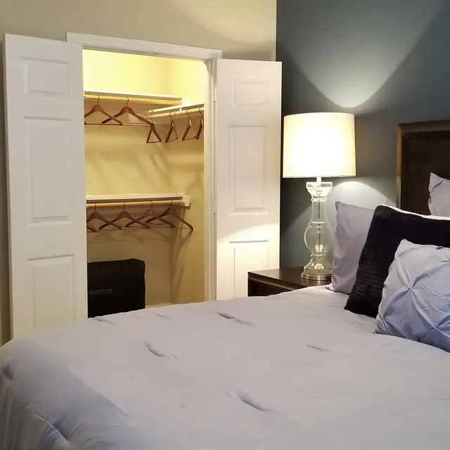 A cozy bedroom featuring a large bed with decorative pillows, a bedside lamp, and a closet. In the background, an open door leads to a bathroom area with a view of a sink and mirror.
