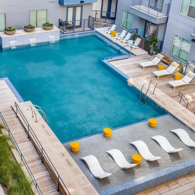 Aerial view of a modern apartment pool area featuring a large, clear swimming pool surrounded by wooden decks and lounge chairs. Yellow planters are placed throughout the area, and there are pathways leading to the poolside.