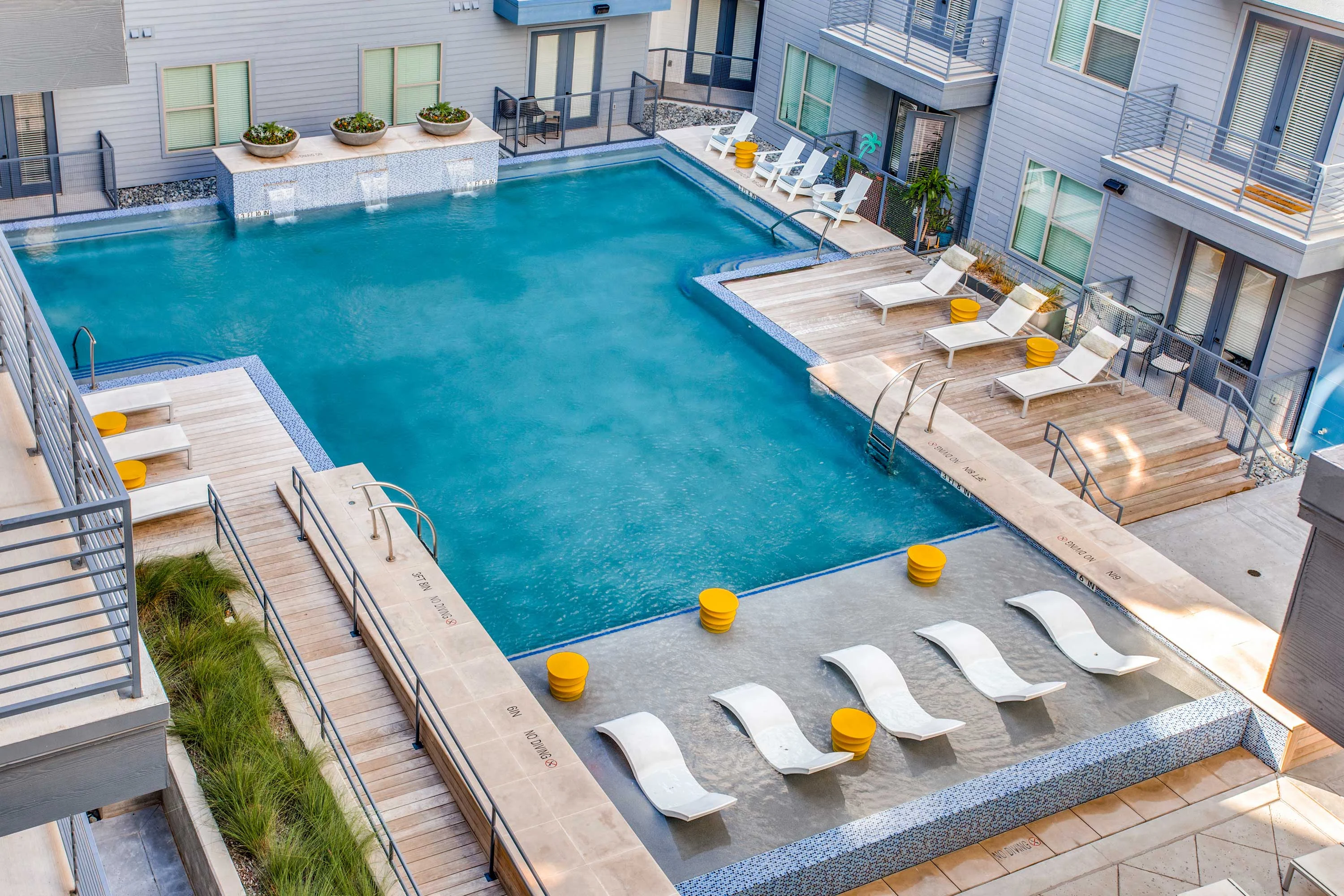 Aerial view of a modern apartment pool area featuring a large, clear swimming pool surrounded by wooden decks and lounge chairs. Yellow planters are placed throughout the area, and there are pathways leading to the poolside.