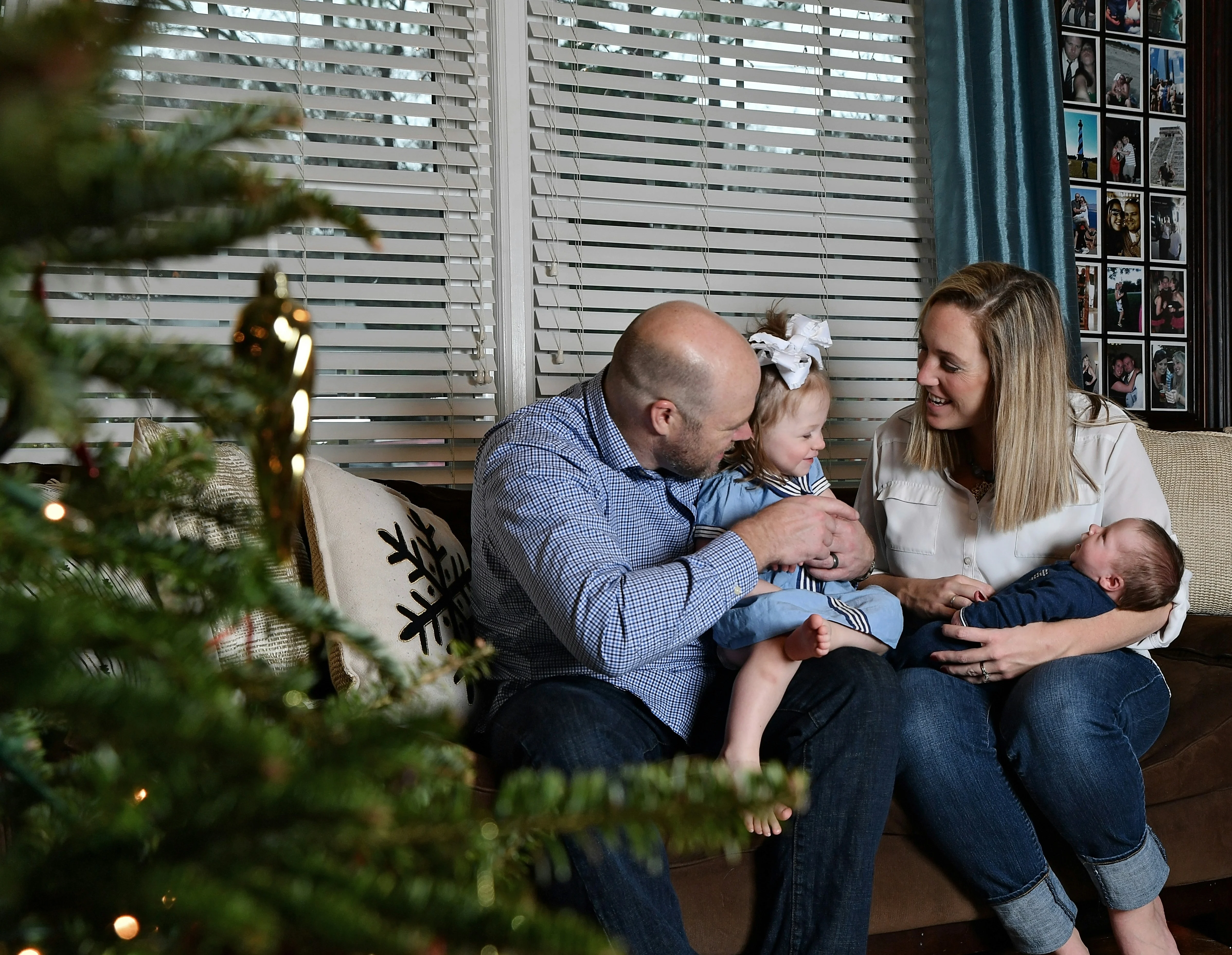 A family of four, including parents and two young children, sitting together on a couch in a cozy living room decorated for the holidays. A Christmas tree is partially visible in the foreground.