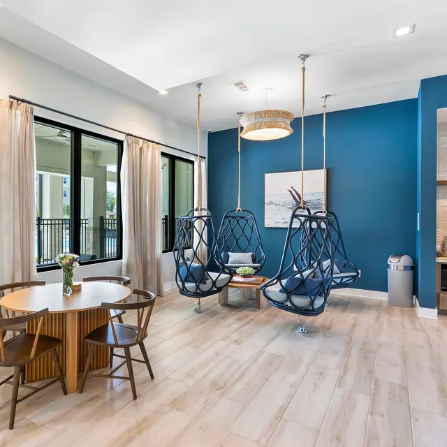 Stylish Living Area with Swing Chairs A modern living area featuring two hanging swing chairs, a round dining table with wooden chairs, and a kitchenette area. The color scheme includes blue and light tones, with large windows and curtains.