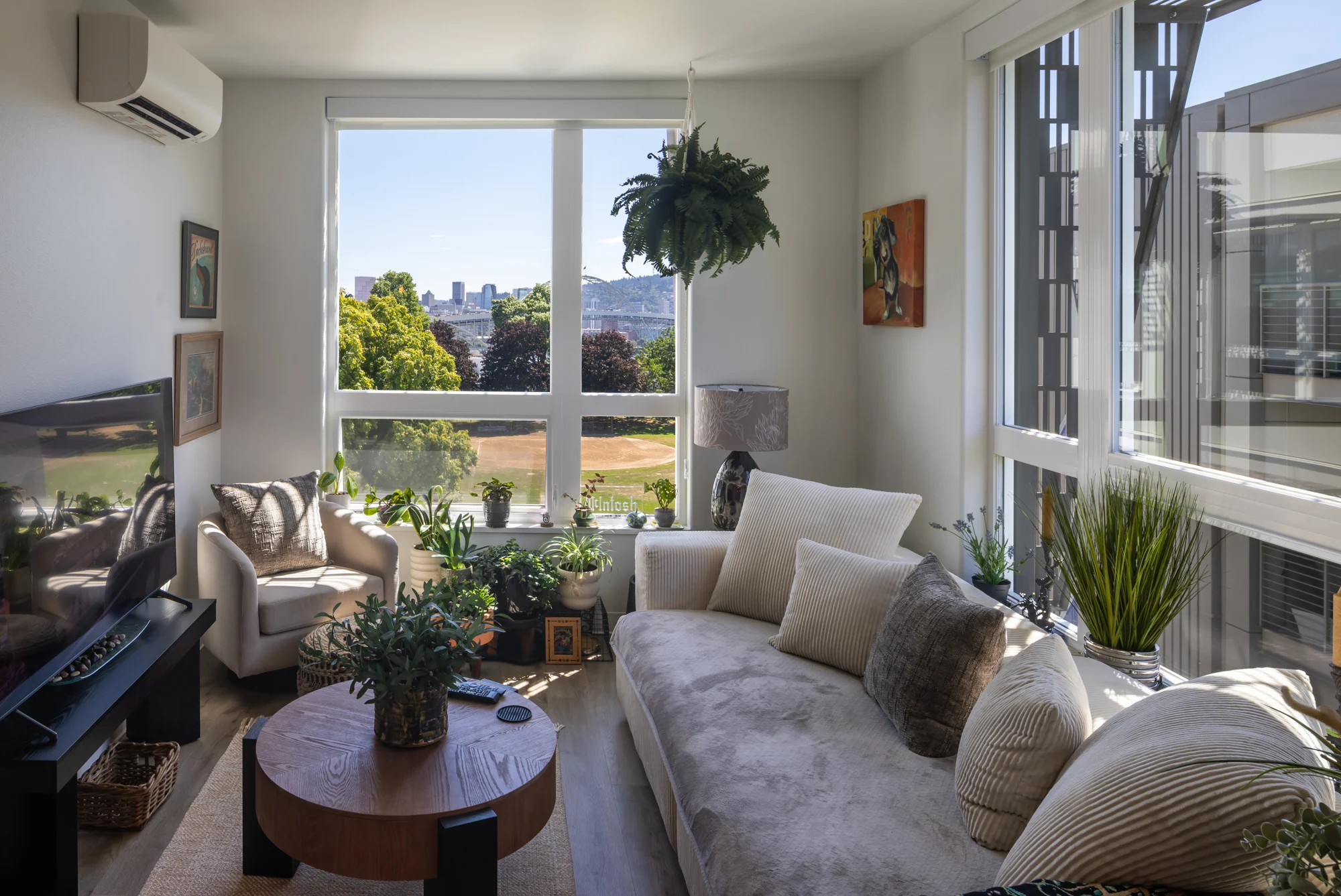 Bright Apartment Living Room A bright living room with a large window showcasing a green landscape. The room features a comfortable beige sectional sofa, a round wooden coffee table, and various indoor plants. There's a small armchair and a TV set against one wall.