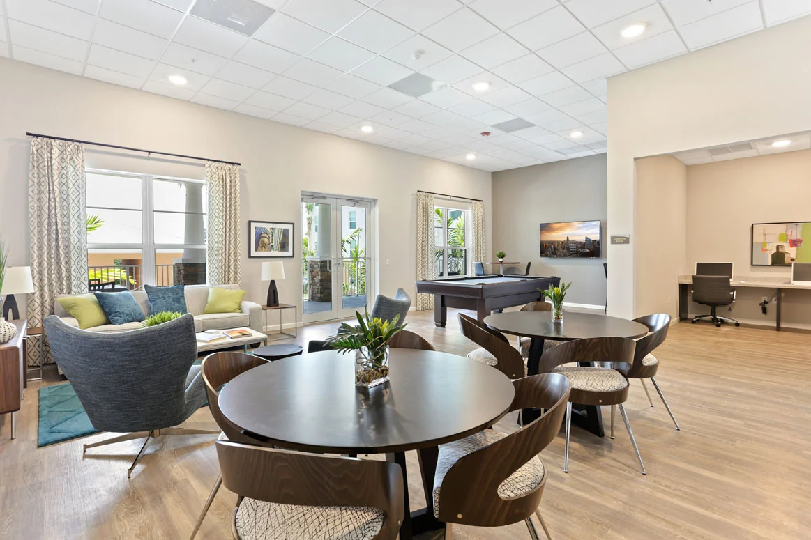 A modern community lounge area with tables and chairs, a sitting area with couches, and large windows providing natural light.
