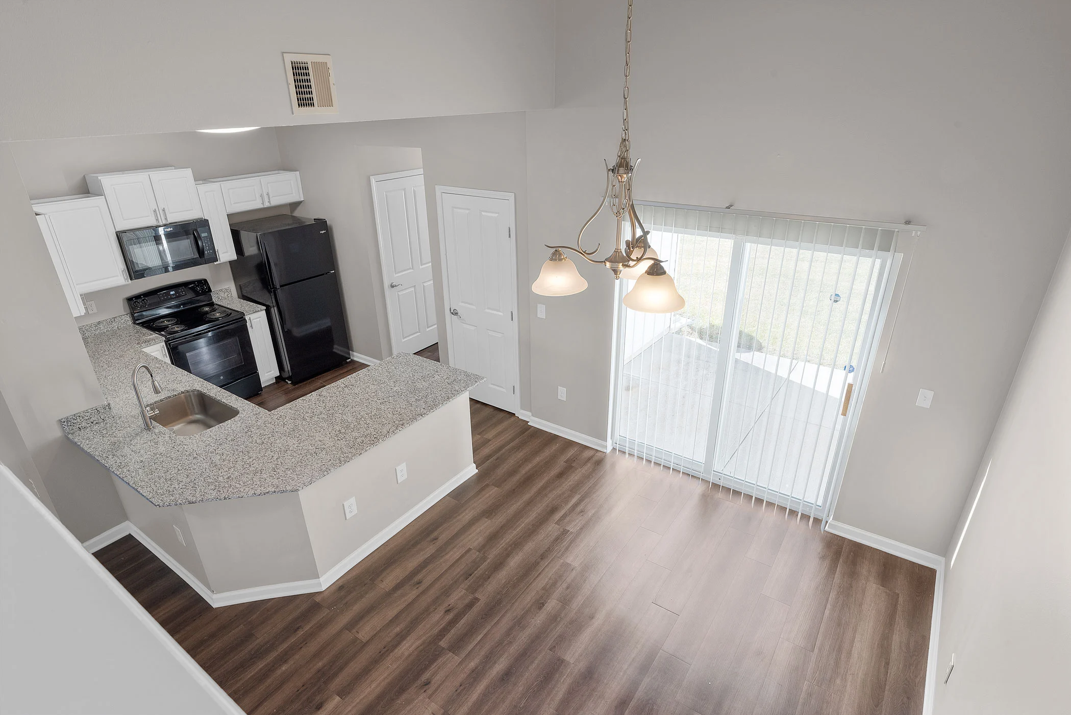 A modern open-concept kitchen and living area featuring a granite countertop, black appliances, hardwood floors, and natural light from sliding glass doors.