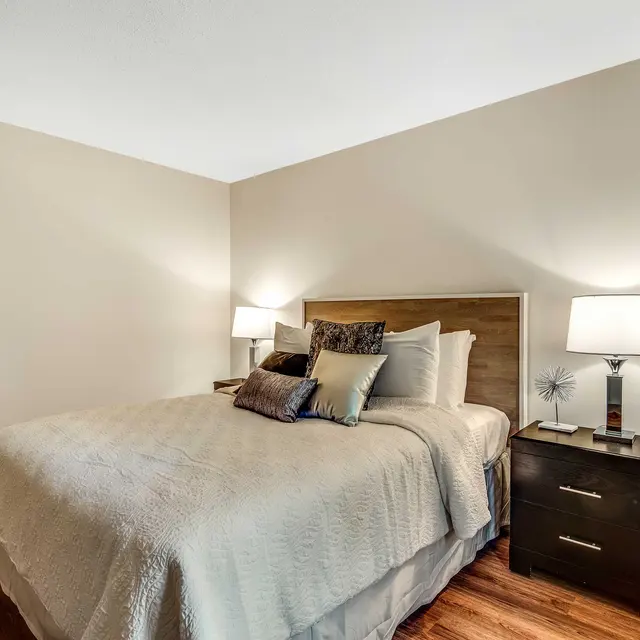 A cozy bedroom featuring a queen-sized bed with a light gray quilt and decorative pillows. There are two bedside lamps on either side of the bed and a dark wood nightstand with a decorative object. The room has warm hardwood flooring and light-colored walls.