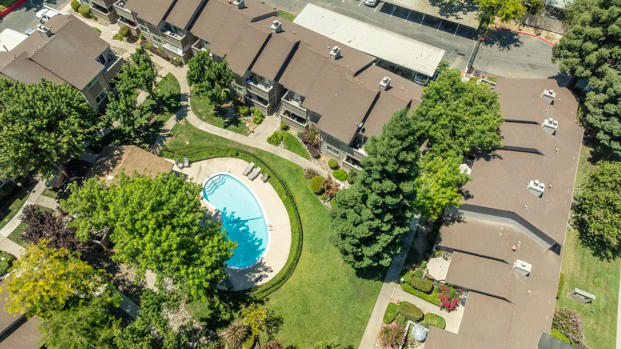 Aerial view of an apartment complex featuring multiple buildings surrounding a circular swimming pool, with vibrant green trees and landscaping throughout the area.