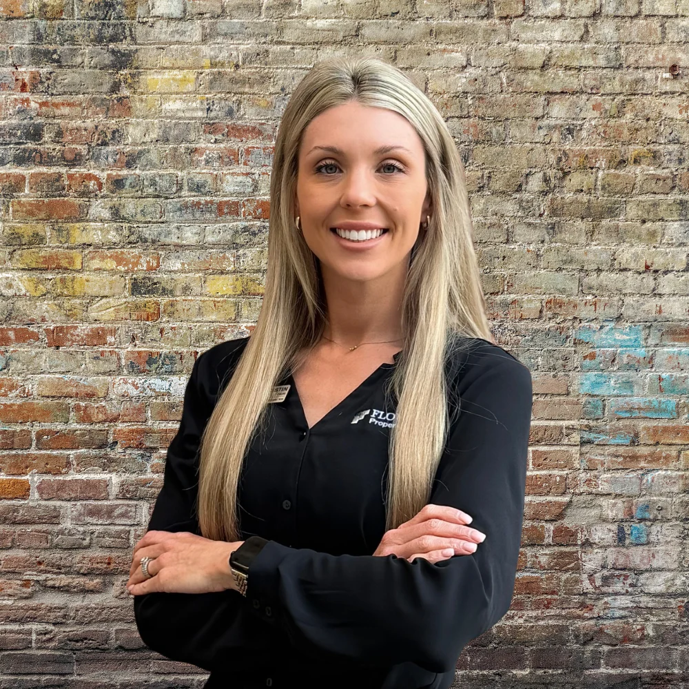 Professional Woman Against Brick Wall A professional portrait of a smiling woman with long, straight hair standing with her arms crossed in front of a brick wall.