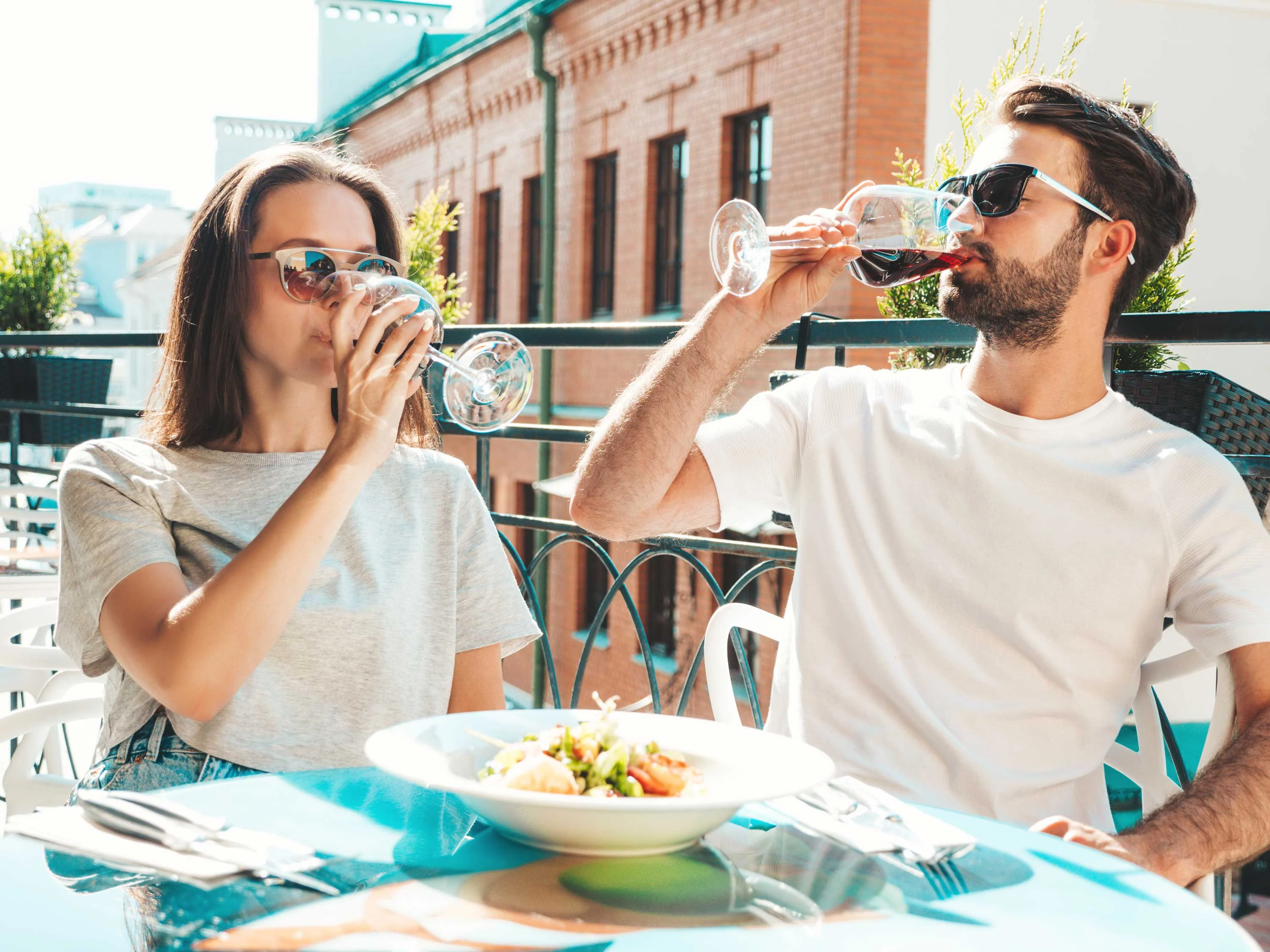 A couple enjoying drinks at an outdoor dining table, dressed casually and wearing sunglasses. In front of them is a plate of salad.