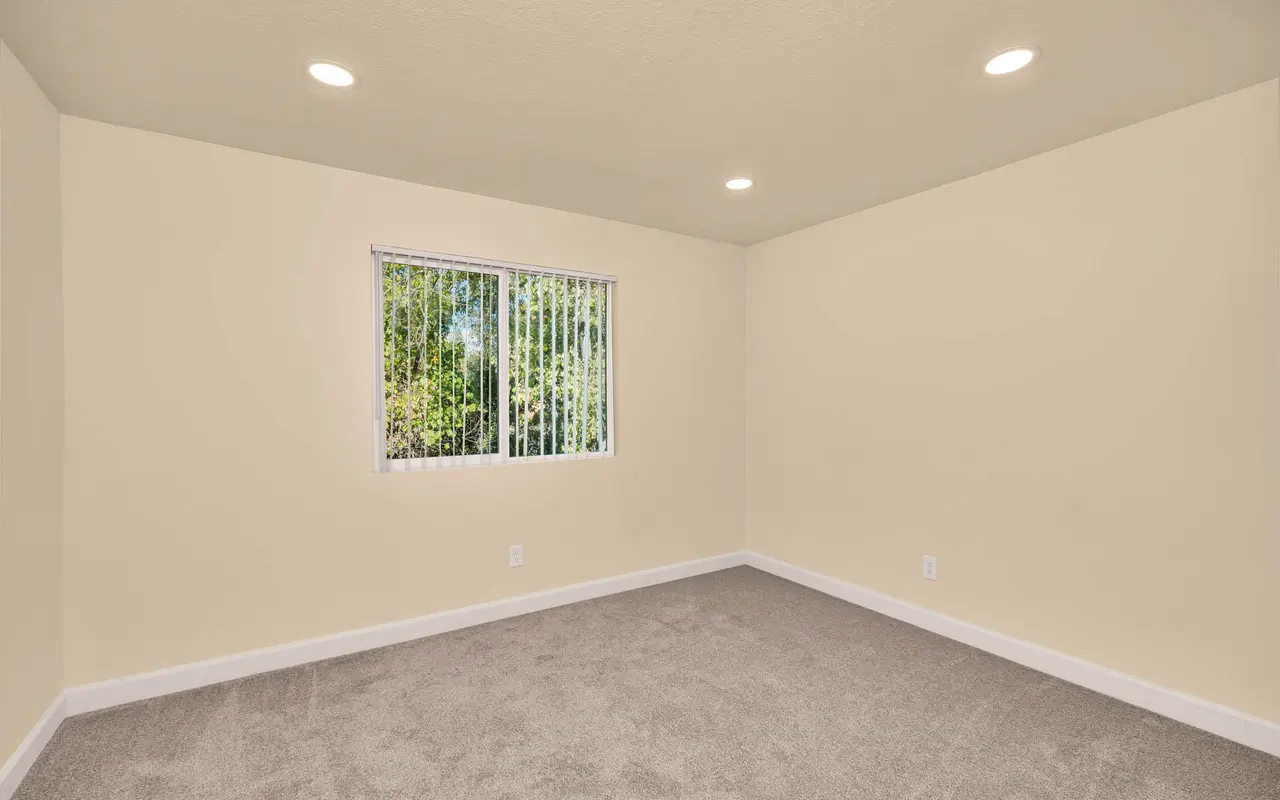 A spacious empty room with light-colored walls, carpeted flooring, and a window with vertical blinds.