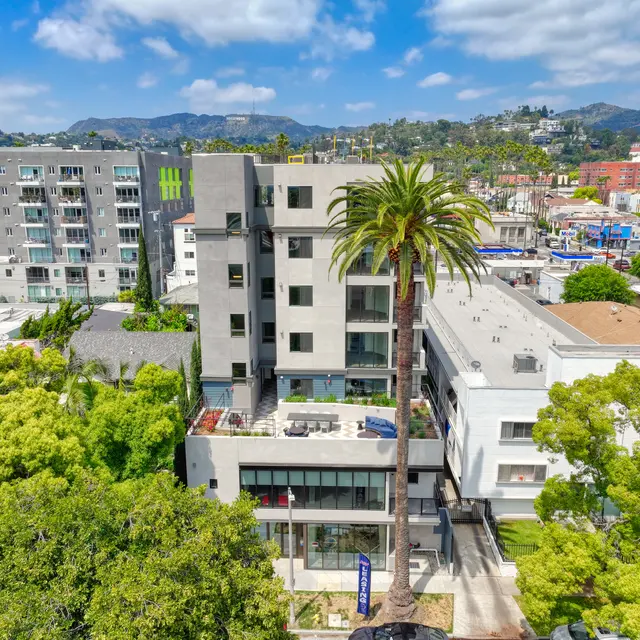 Modern Apartment Building with Lush Surroundings A modern multi-story apartment building surrounded by palm trees and greenery, with a view of the hills in the background under a partly cloudy sky.