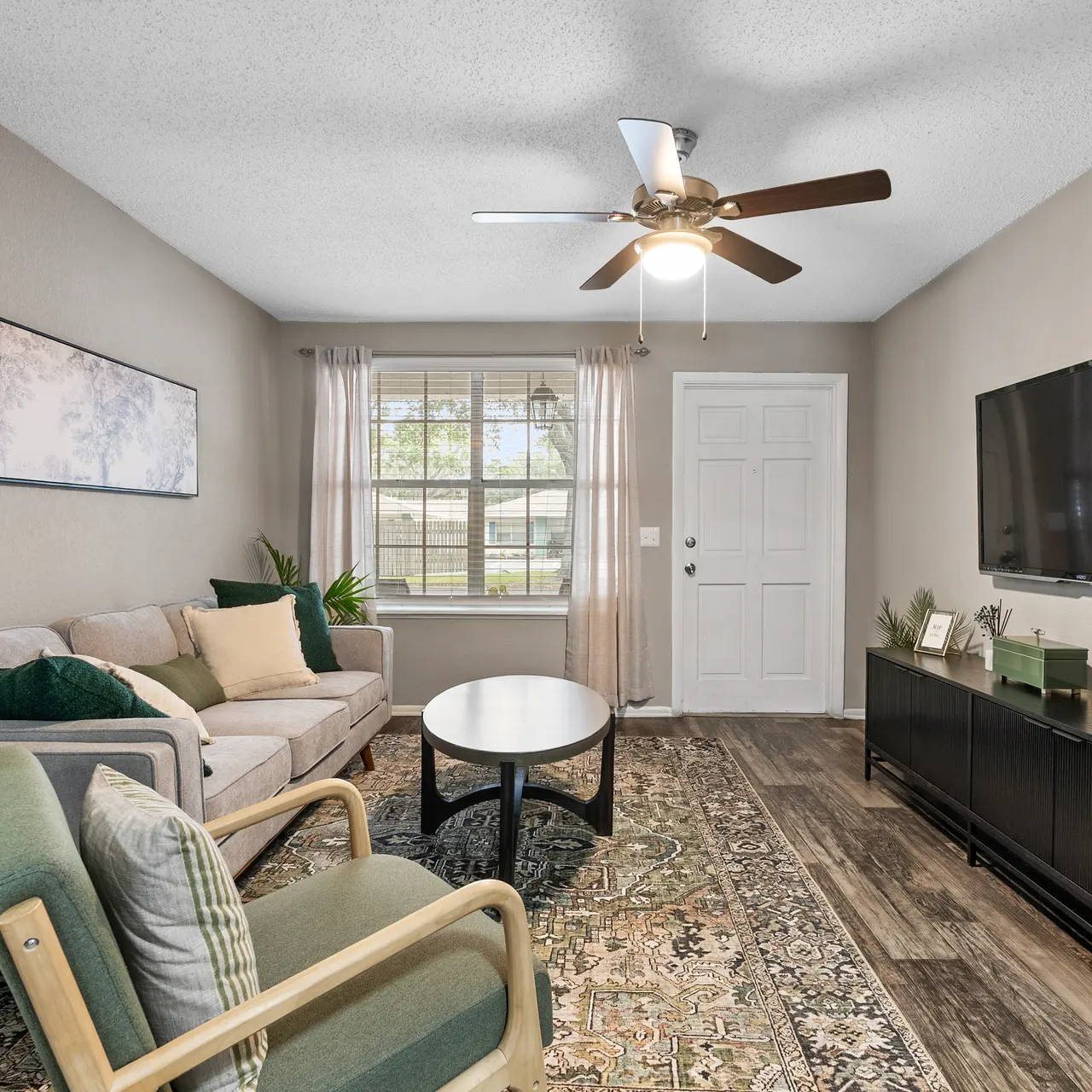 A cozy living room featuring a sofa, green accent chairs, a round coffee table, and a television mounted on the wall. Natural light streams in through a window with white curtains.