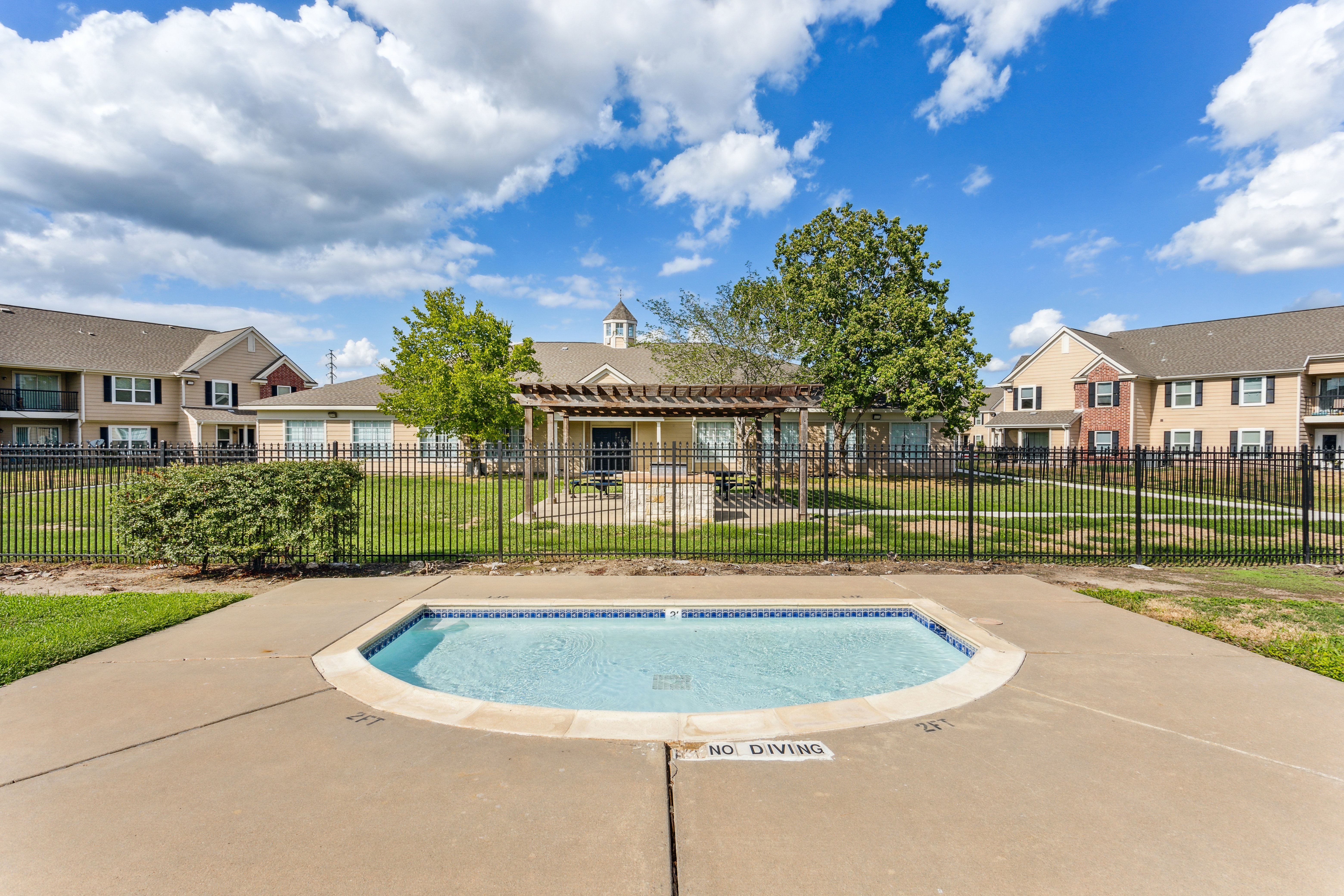 Small hot tub with a no diving sign in an apartment community area surrounded by green grass and a fence, with several buildings in the background under a partly cloudy sky.