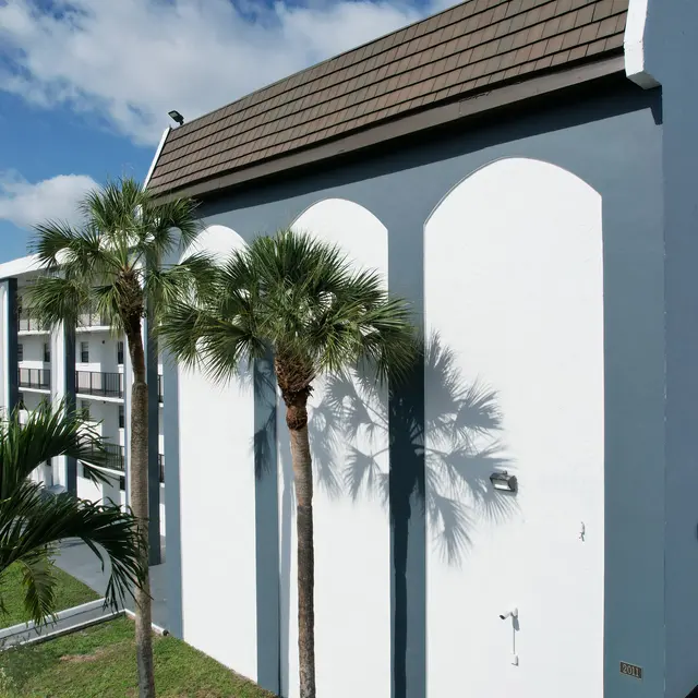 A modern multi-story building with white walls and a brown sloped roof, flanked by palm trees. The blue sky is visible overhead with fluffy clouds.