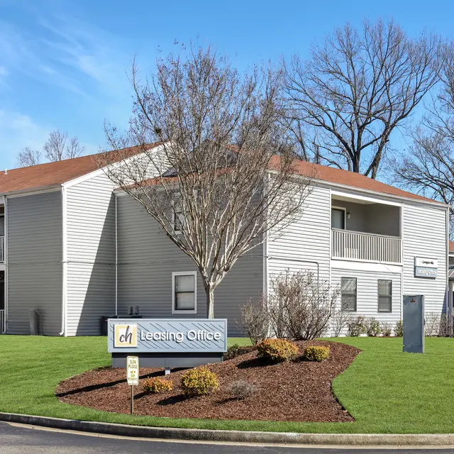 Exterior view of a leasing office building with a sign in front, surrounded by green grass and trees, under a clear blue sky.