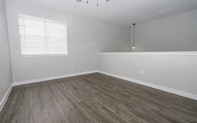 A spacious empty room with light gray walls and modern laminate flooring. There is a window with white blinds allowing natural light, and a small pendant light is hanging from the ceiling. A half wall separates this area from a neighboring space, creating an open feel.
