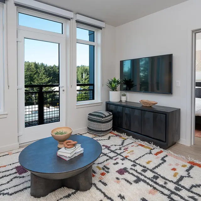 A bright and modern living room featuring large windows with a view of greenery, a round coffee table, a cozy blue sofa, and a dark TV cabinet. The floors are light wood, and the rug has a colorful geometric design. A doorway leads to a bedroom with a bed visible in the background.