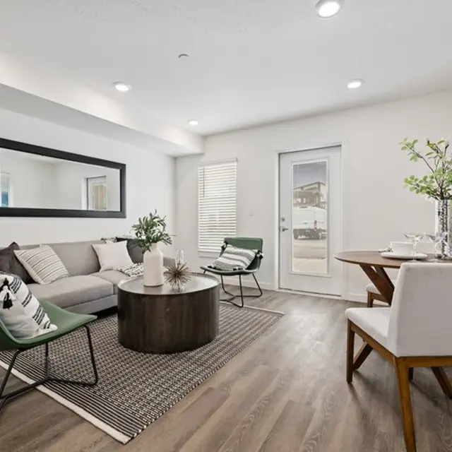 A modern living room featuring a grey sofa with assorted cushions, a round coffee table, and a striped rug. There are two chairs, one green and one white, alongside a small dining table with tableware. Large windows let in natural light.