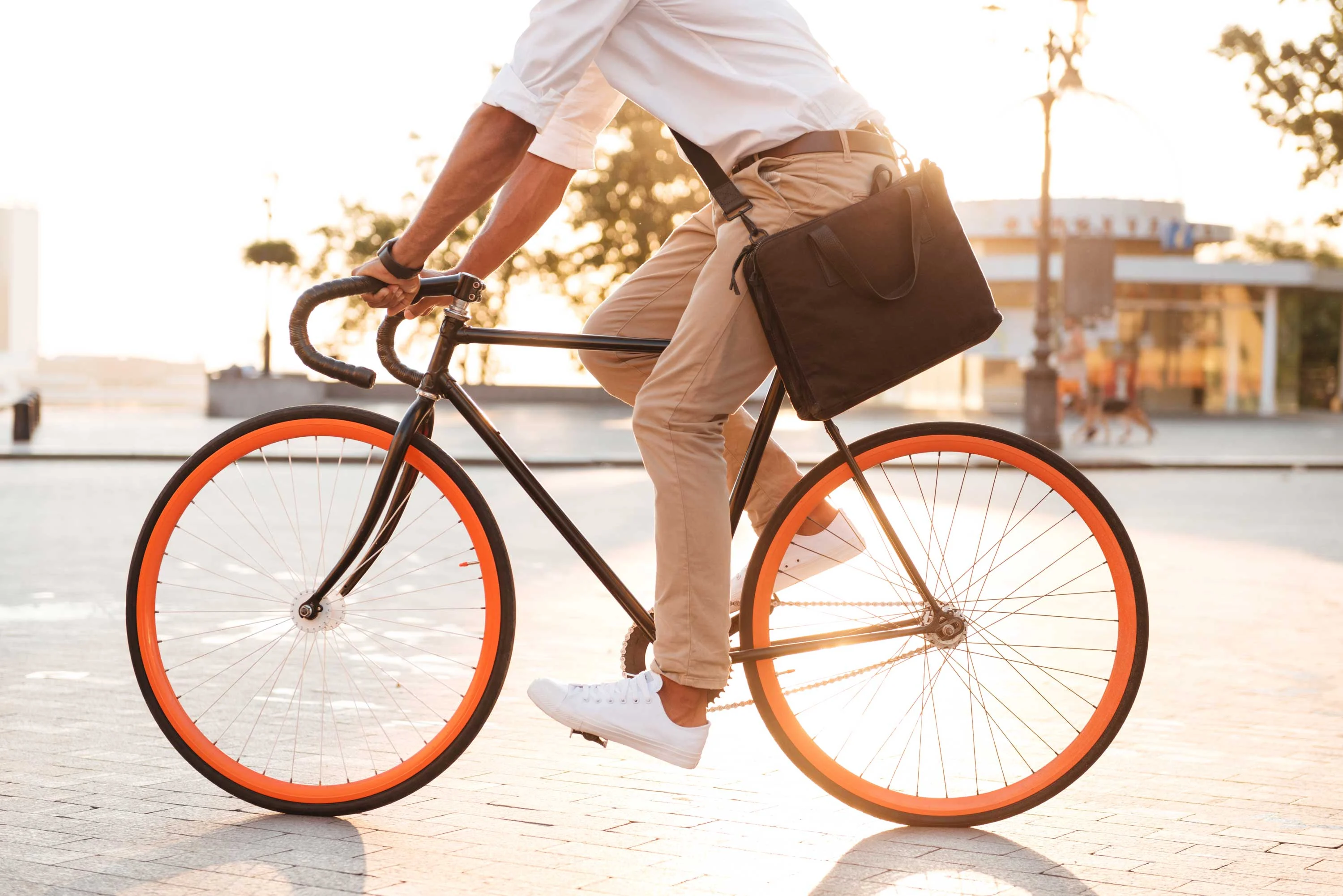 Cyclist in Urban Landscape A person riding a bicycle with orange wheels, wearing a white shirt and beige pants, carrying a black bag over their shoulder, in a sunny outdoor setting.