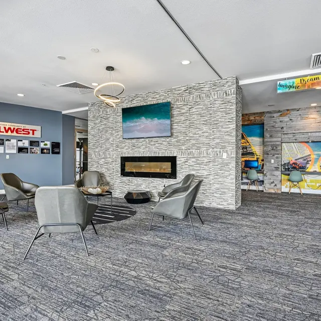 Modern Community Lounge Area A modern community lounge area with several gray chairs arranged around a central rug. The room features a stone wall with a fireplace and a television mounted above it, along with colorful wall art depicting a playful scene.