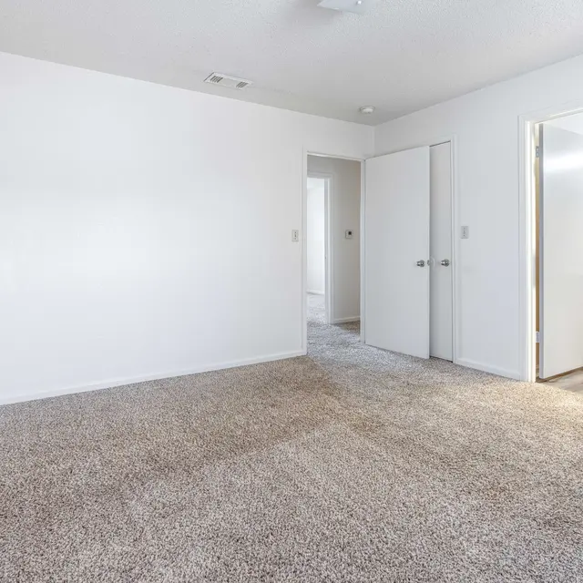 A spacious empty room with light-colored walls and carpet flooring, featuring a doorway leading to another room and a bathroom in the background.