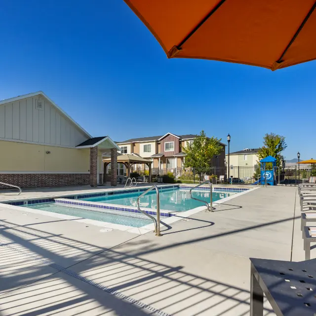 Relaxing Pool Area A clean swimming pool area with chaise lounges and an orange umbrella providing shade, surrounded by a fence and colorful landscaping in the background.
