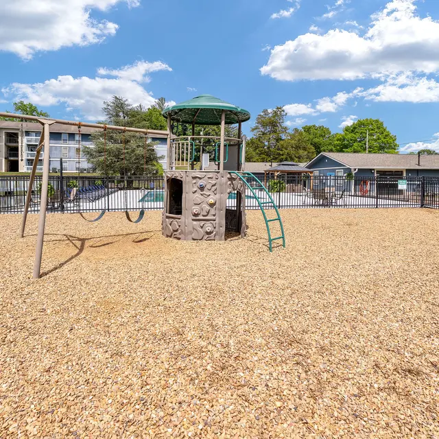 A sunny playground featuring a climbing structure and swings, surrounded by gravel and a safety fence, with residential buildings in the background.