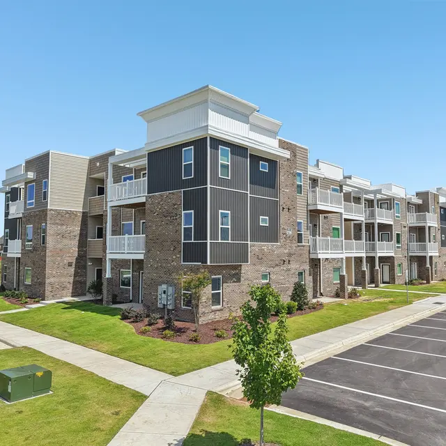 A modern apartment complex featuring multiple three-story buildings with a mix of dark and light exterior finishes. The area is landscaped with grass and small trees, and there is a paved parking lot in the foreground.