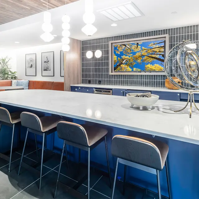 Resident Lounge Kitchen A modern kitchen area featuring a long, light-colored island with blue accents and bar stools. There are decorative items on the island and artwork on the wall, with pendant lighting above. The space is designed with a contemporary aesthetic.