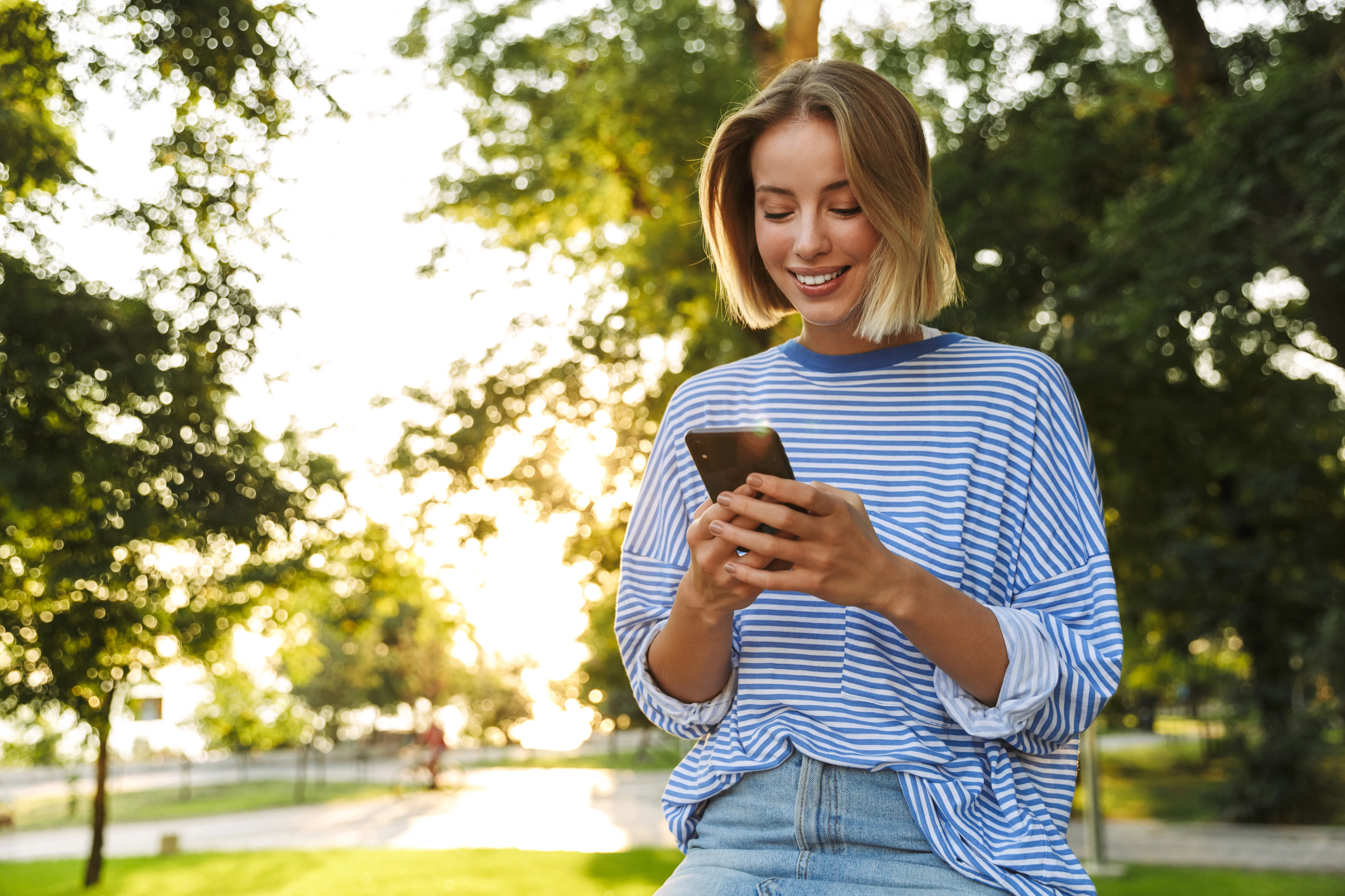 A young woman with short blonde hair wearing a blue and white striped shirt sits on a bench in a park, smiling at her smartphone with trees and sunlight in the background.