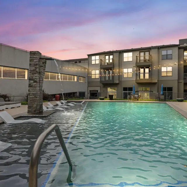 A modern apartment complex featuring a beautiful swimming pool surrounded by lounge chairs and a stone water feature, set under a colorful sunset sky.