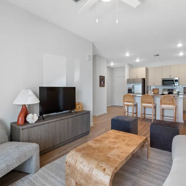 A modern living room featuring a light grey couch, a wooden coffee table, and a small flat-screen TV on a dark cabinet. The kitchen is visible in the background with beige cabinets and high stools at a counter.