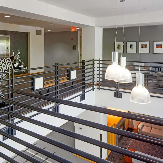 A modern interior space with multiple levels, featuring a balcony with horizontal railings, pendant lights hanging above, and abstract art on the walls. A contemporary patterned couch is visible in the background.