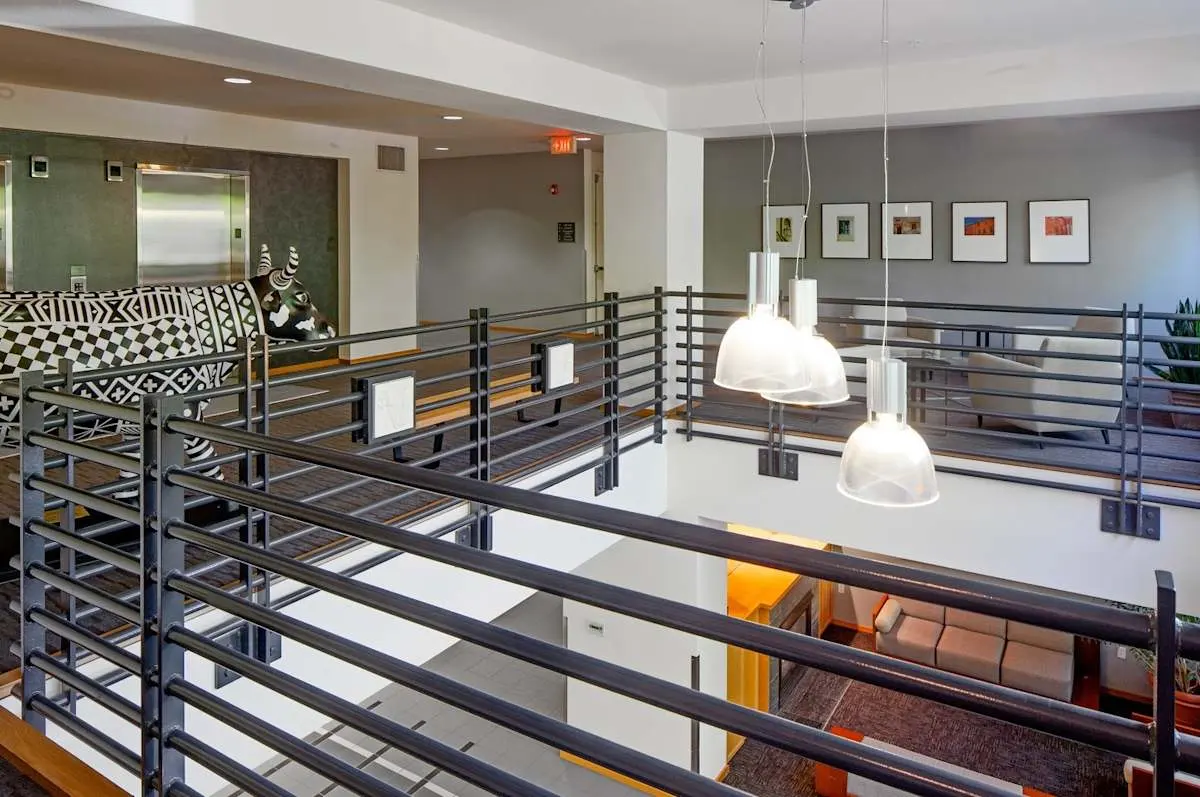 A modern interior space with multiple levels, featuring a balcony with horizontal railings, pendant lights hanging above, and abstract art on the walls. A contemporary patterned couch is visible in the background.