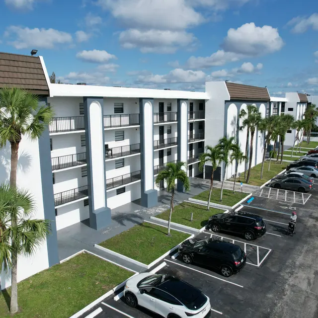A modern multi-story apartment complex viewed from above, featuring palm trees, balconies, and a parking lot filled with cars.