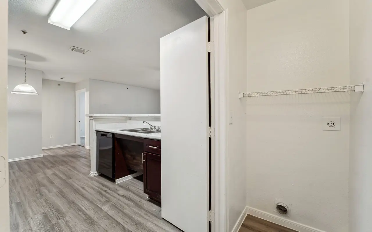 A view of an interior space with a kitchen area visible through an open doorway. There are light brown wooden floors, white walls, and a simple light fixture. A closet or pantry with a wire shelf is also visible.