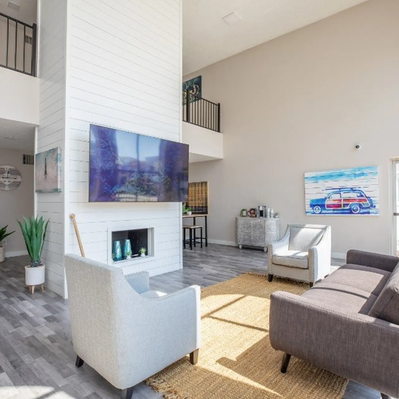 A spacious and modern living room featuring a tall ceiling, large windows, and a flat-screen TV mounted on a white shiplap wall. Two armchairs and a dark sofa are arranged around a light-colored area rug, with a small potted plant nearby. There's a painting of a classic car on the wall and an entrance door visible at the side.