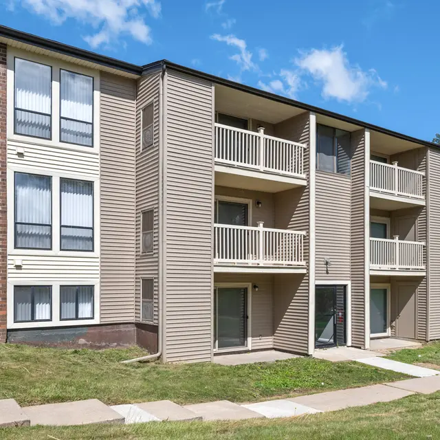 A three-story apartment building with a combination of brick and vinyl siding. The building has multiple balconies and a grassy area in front. The sky is partly cloudy.