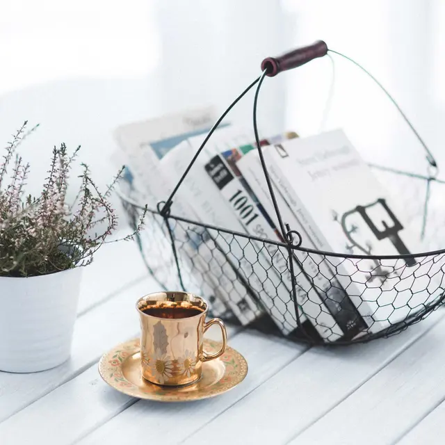 A cozy table setting featuring a wire basket filled with magazines, a small potted plant, and a gold coffee cup on a saucer.